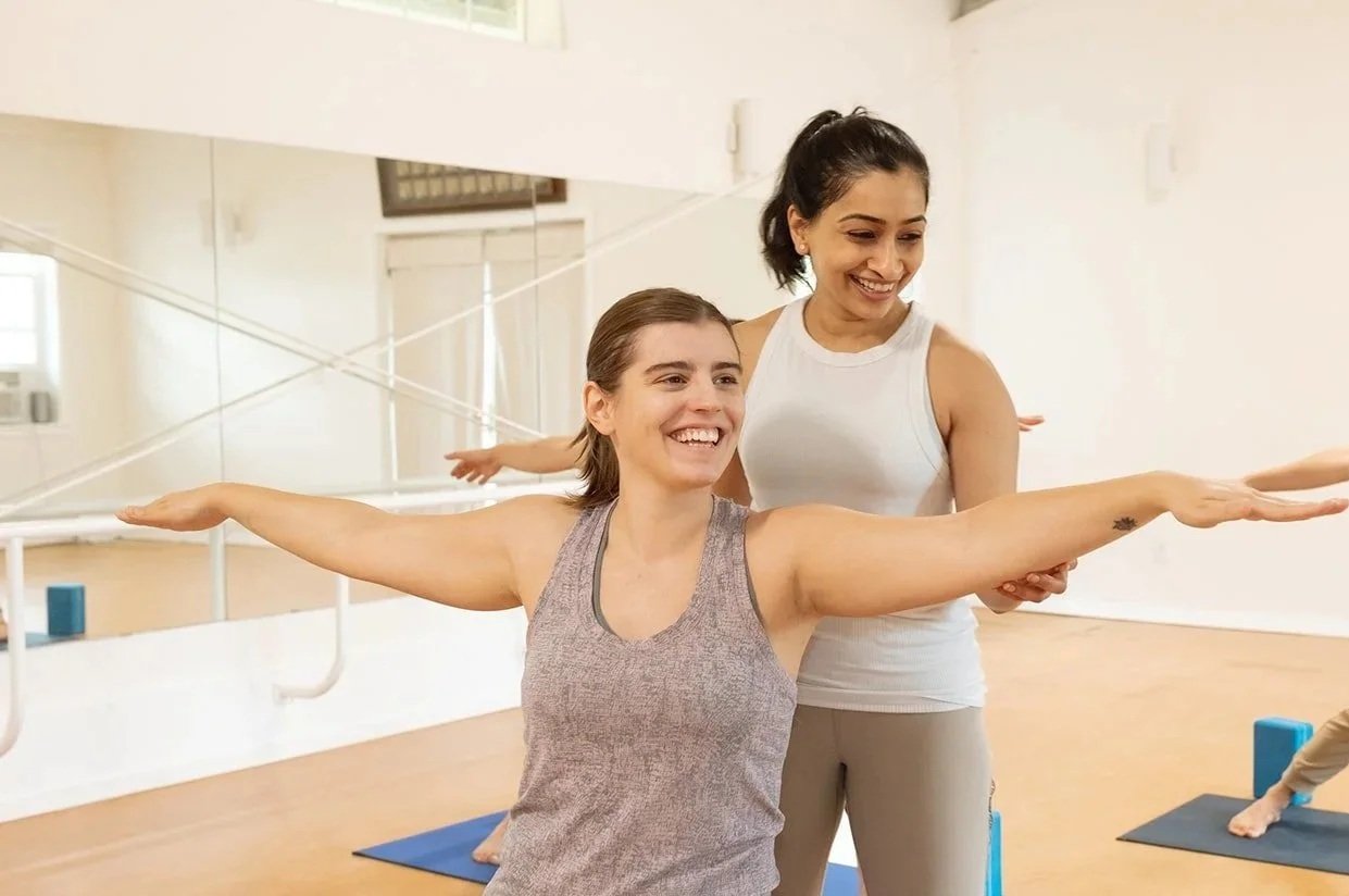 Arundhati smiling behind a new student who stands in a warrior pose with arms wide, gently guiding her posture.